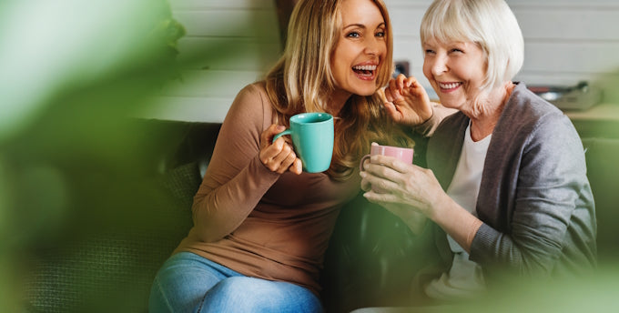 Happy senior mother with adult daughter sitting on couch and holding cups with coffee or tea at home.